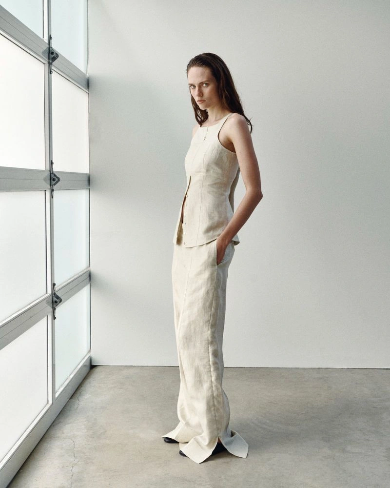 Model wearing a cream linen sleeveless vest and matching wide-leg trousers standing in a minimalist white room.