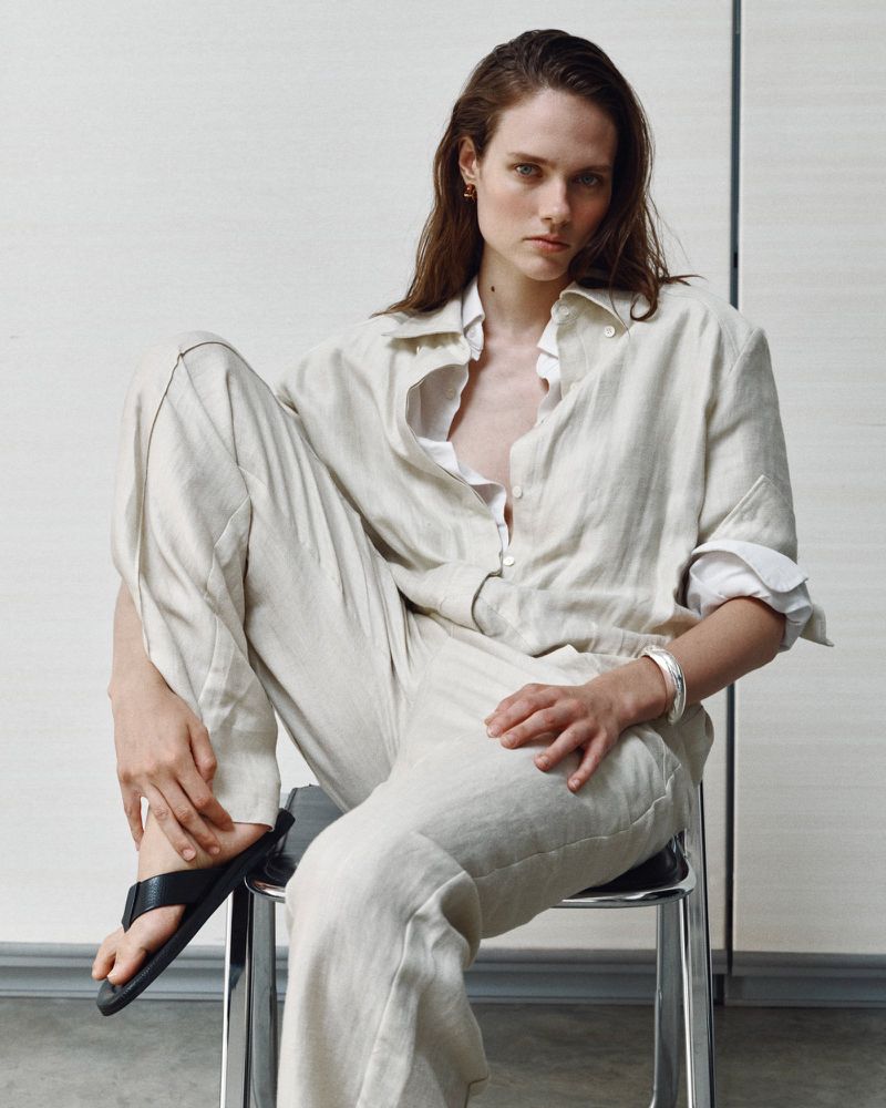 Model Kate McNamara seated on a metal chair wearing neutral linen trousers and a matching button-down shirt for ACCORDA SS27