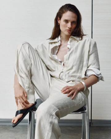 Model Kate McNamara seated on a metal chair wearing neutral linen trousers and a matching button-down shirt for ACCORDA SS27