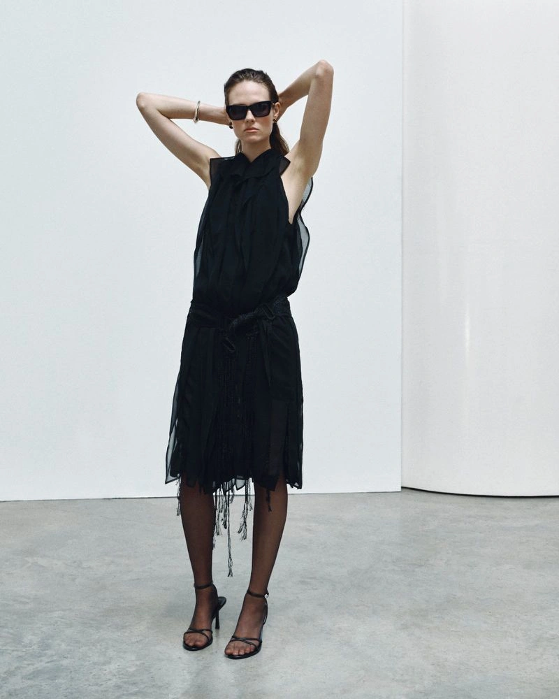 Model wearing a black dress and sunglasses with hands behind head against a plain white studio background