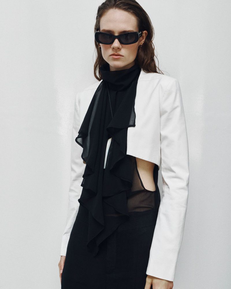 Model wearing dark sunglasses, a black sheer top, and a white cropped jacket against a plain white studio background.