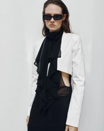 Model wearing dark sunglasses, a black sheer top, and a white cropped jacket against a plain white studio background.