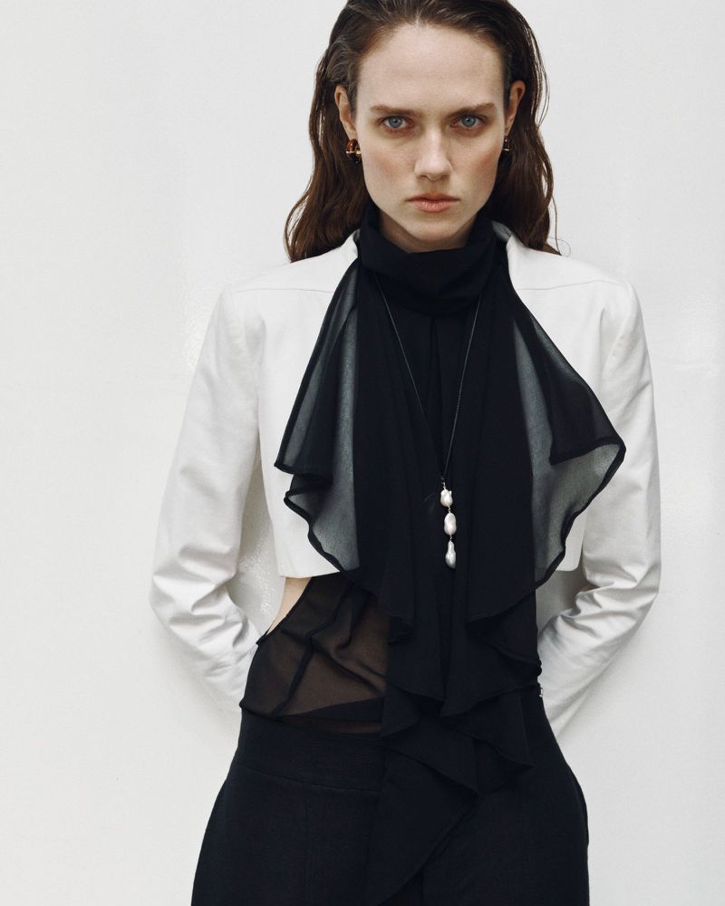 Model in white cropped blazer and black sheer layered top against a plain white studio background