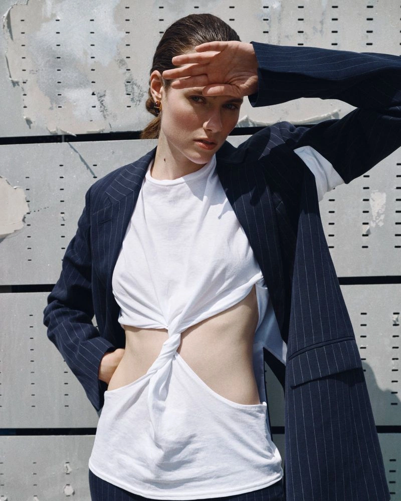 Model wearing a navy pinstripe blazer and white cut-out top posing against an industrial metal panel background.