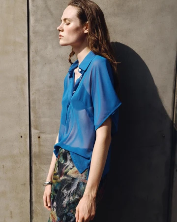 Model wearing a blue sheer short-sleeve button-up shirt and patterned skirt standing in profile against a textured grey concrete wall.