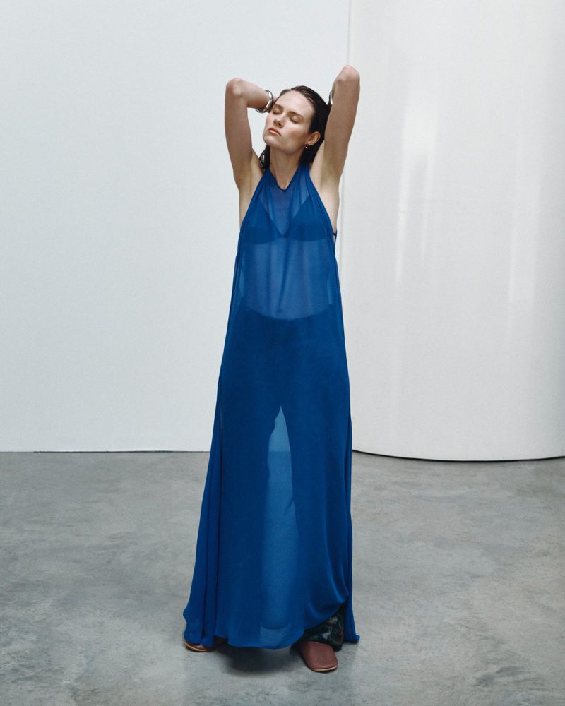 Model wearing a long blue sheer slip dress posing with arms raised in a minimalist studio setting.