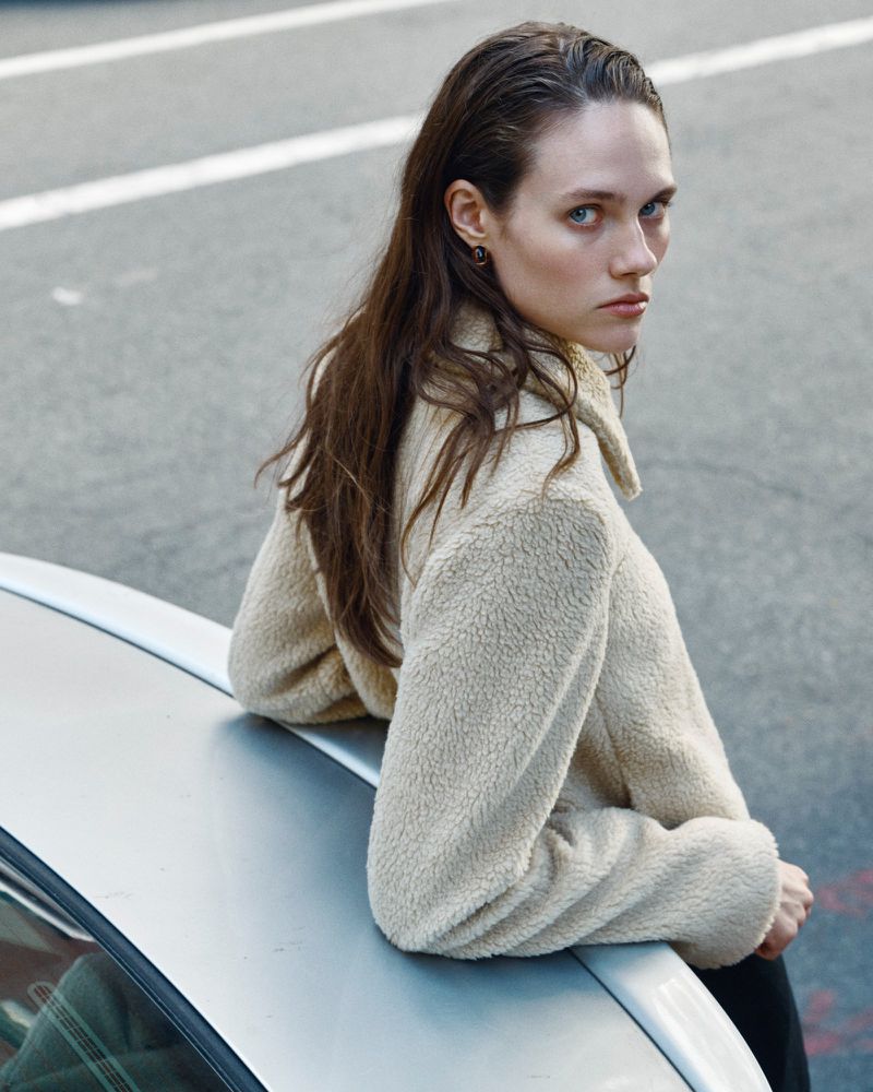 Model in cream textured jacket leaning against a silver car on asphalt road