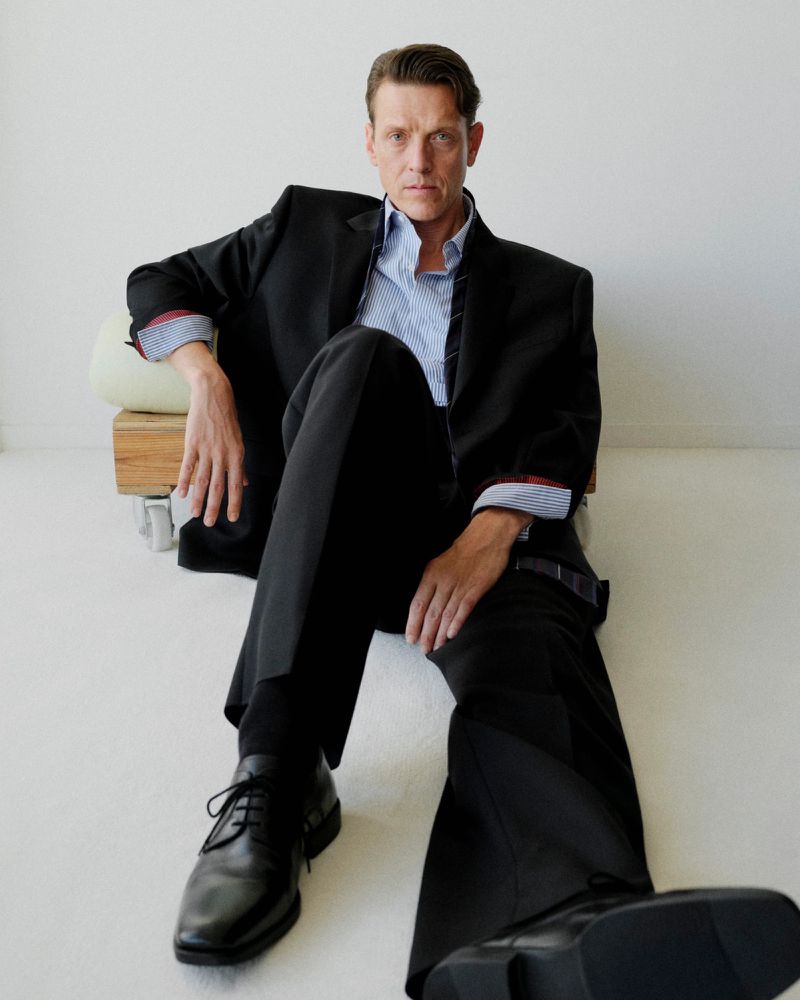 Model seated on the floor wearing a black suit and blue striped shirt against a plain white background