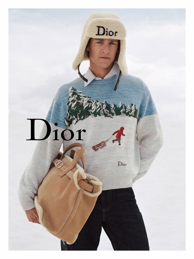 Model wearing a winter sweater and plush Dior trapper hat holding a tan leather bag against an overcast sky
