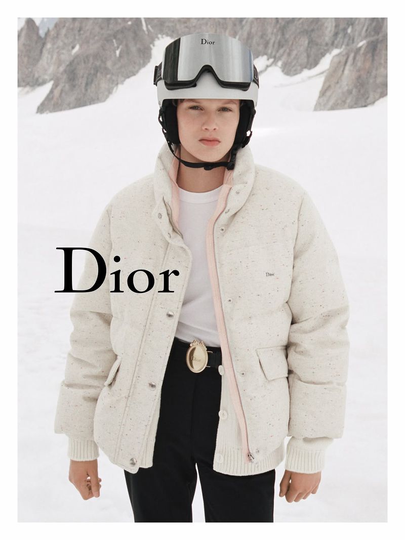 Model in white ski helmet and cream puffer jacket standing before snowy mountain landscape for Dior Alps campaign.