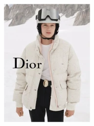 Model in white ski helmet and cream puffer jacket standing before snowy mountain landscape for Dior Alps campaign.