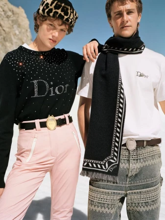 Two models wearing Dior winter apparel outdoors under a clear blue sky, featuring pink ski trousers and patterned trousers.