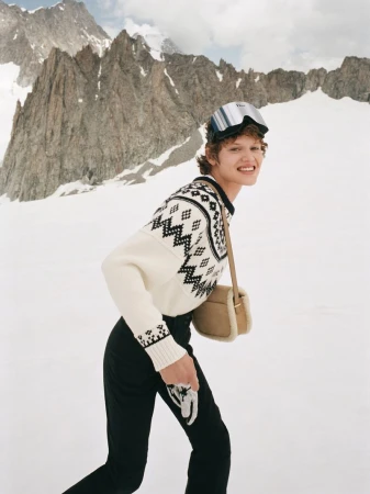 Model wearing patterned knit sweater and ski goggles with a crossbody bag against snowy mountain backdrop