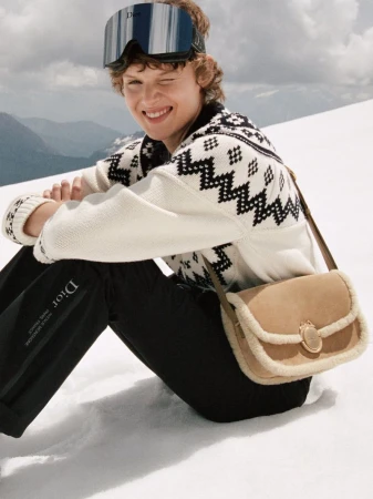 Model wearing mirrored ski goggles and a patterned white sweater sitting on a snowy mountain slope with a tan shearling handbag