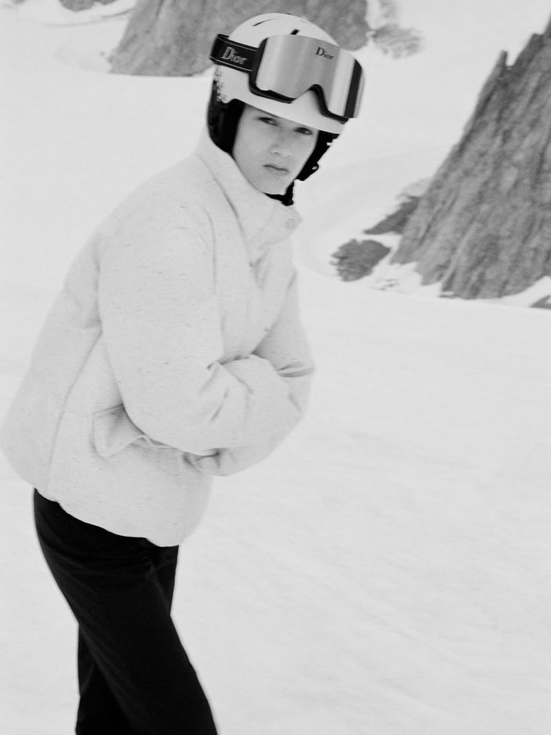 Model wearing a white ski jacket and helmet with goggles in a snowy mountain landscape