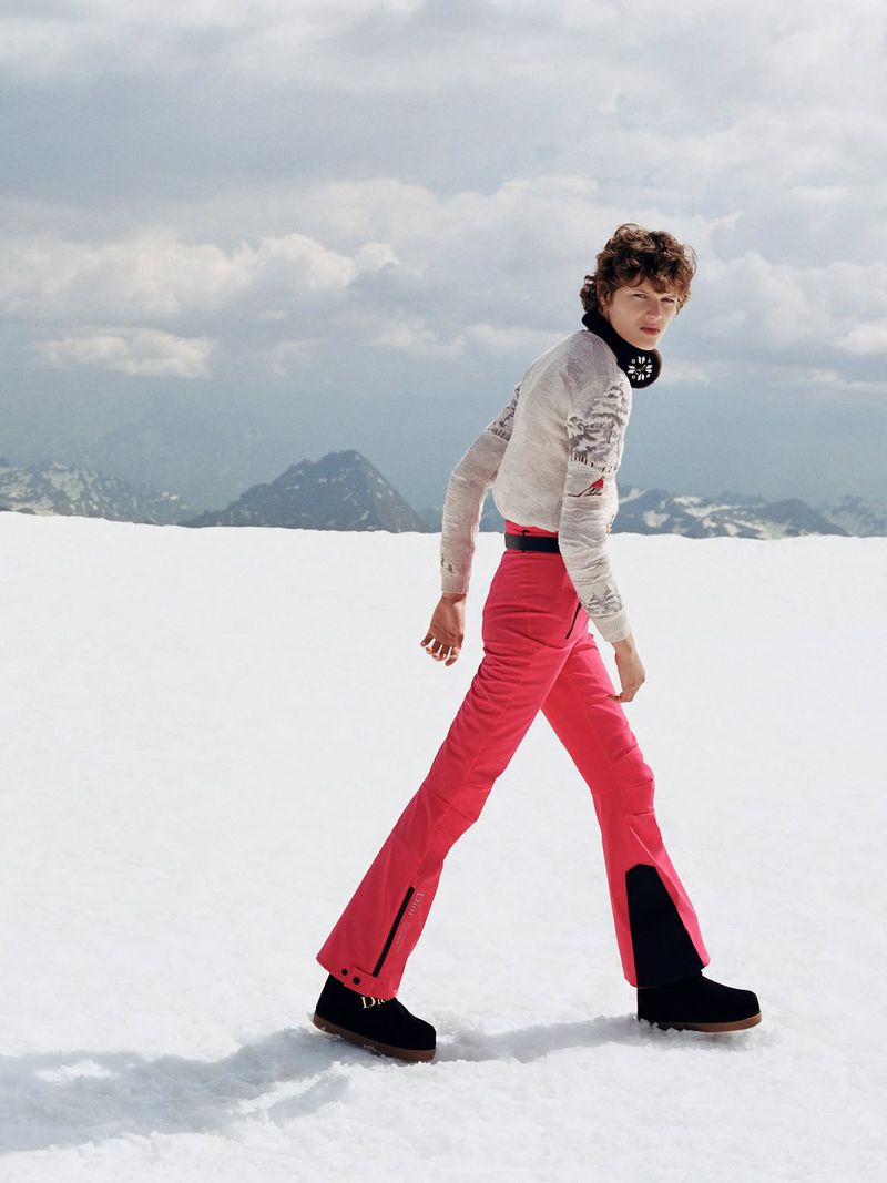 Model wearing patterned sweater and pink ski pants walking across a snowy mountain landscape under an overcast sky.