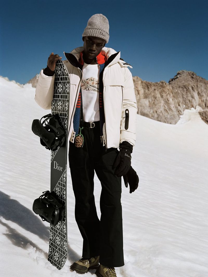 Model holding a snowboard wearing a white puffer jacket, knit beanie, and black ski pants against a snowy mountain backdrop.