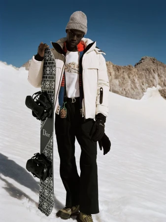 Model holding a snowboard wearing a white puffer jacket, knit beanie, and black ski pants against a snowy mountain backdrop.