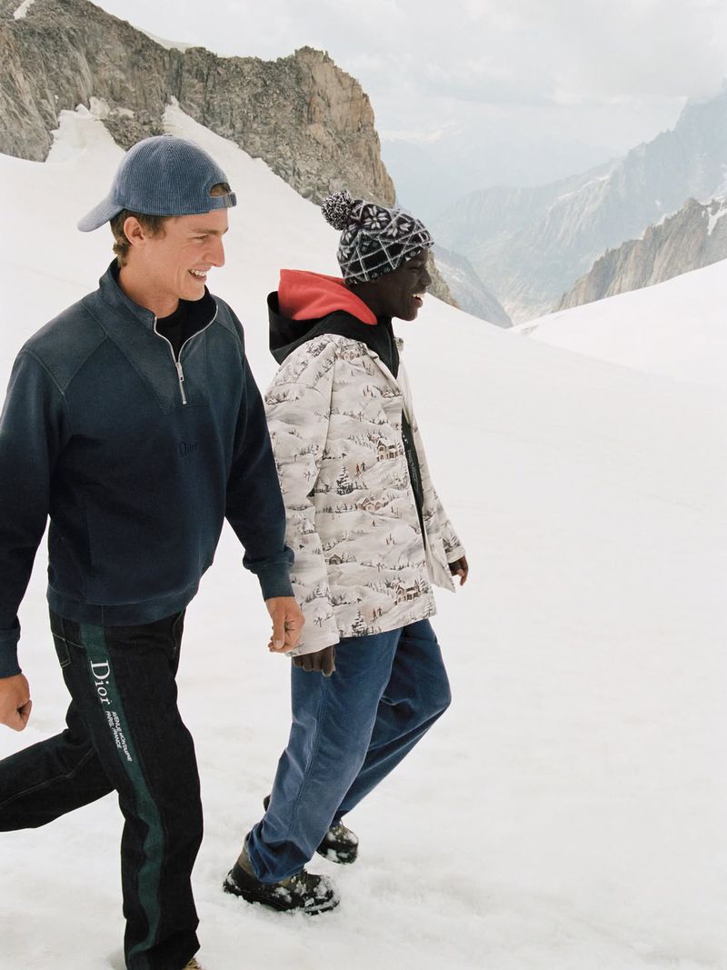 Two models walking outdoors on a snowy mountain slope wearing winter apparel including a patterned jacket and baseball cap