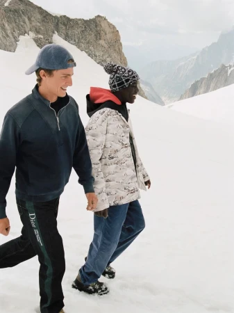 Two models walking outdoors on a snowy mountain slope wearing winter apparel including a patterned jacket and baseball cap