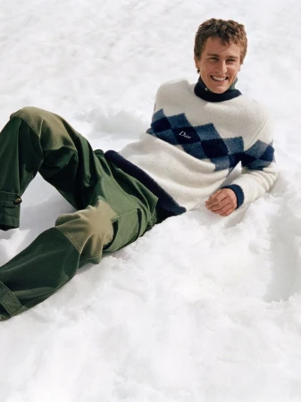 A man smiling while reclining in the snow, wearing an argyle sweater and green trousers for the Dior Alps 2026 campaign.