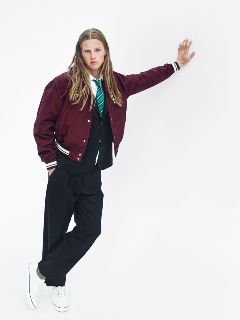 A young male model with long blonde hair leaning against a plain white wall, wearing a burgundy bomber jacket over a dark vest, white shirt, green striped tie, dark trousers, and white sneakers.