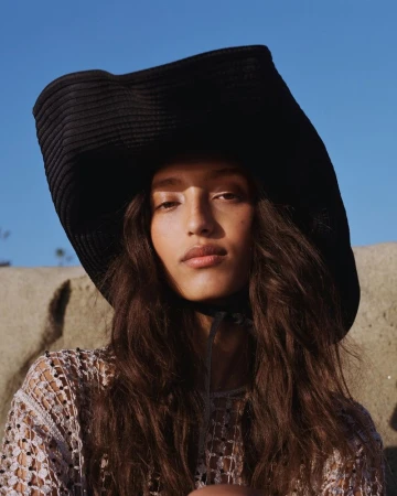 Model wearing a large black sun hat and open-weave crochet top against a clear blue sky and rocky terrain background.