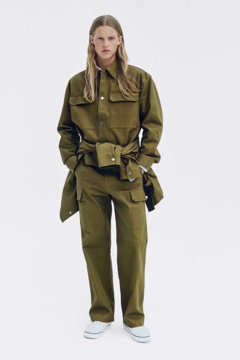 Model with long blonde hair wearing an olive green utility shirt and matching pants against a plain white background