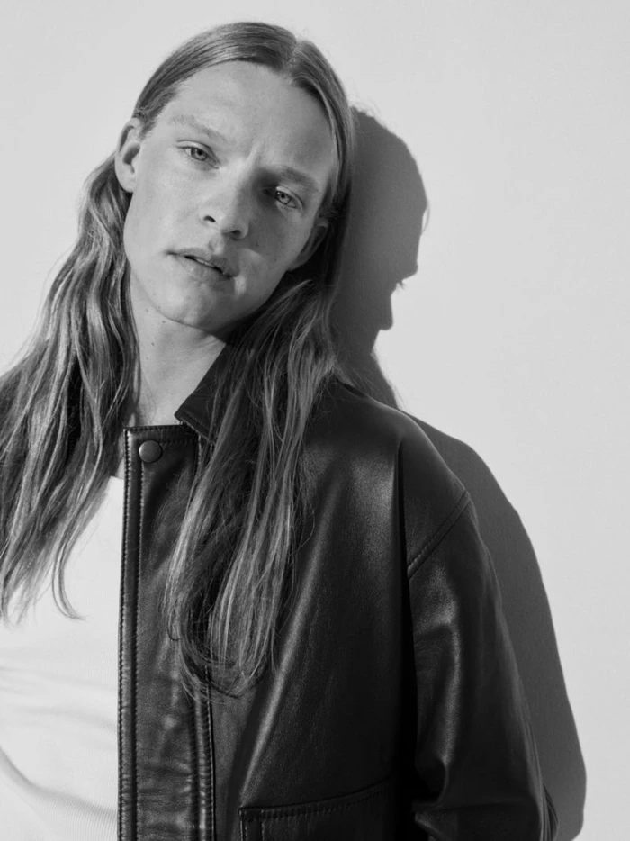 Model with long blonde hair wearing a dark leather jacket against a light studio wall with cast shadow