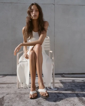 Model sitting on a white chair wearing a white ribbed dress against an outdoor concrete wall