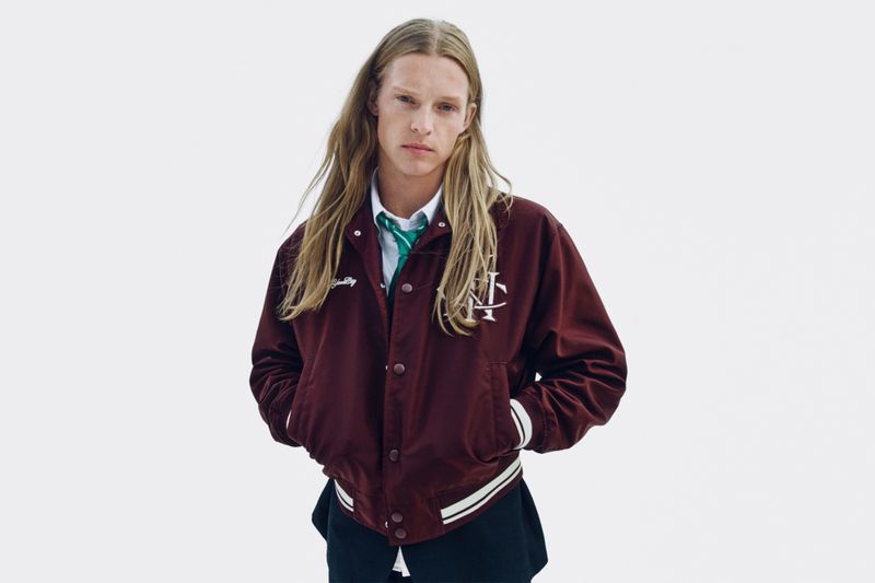 Model with long blonde hair wearing a maroon bomber jacket and green tie against a white background