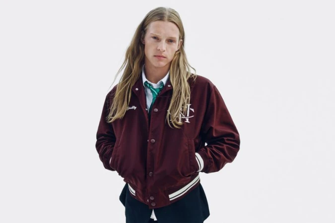 Model with long blonde hair wearing a maroon bomber jacket and green tie against a white background
