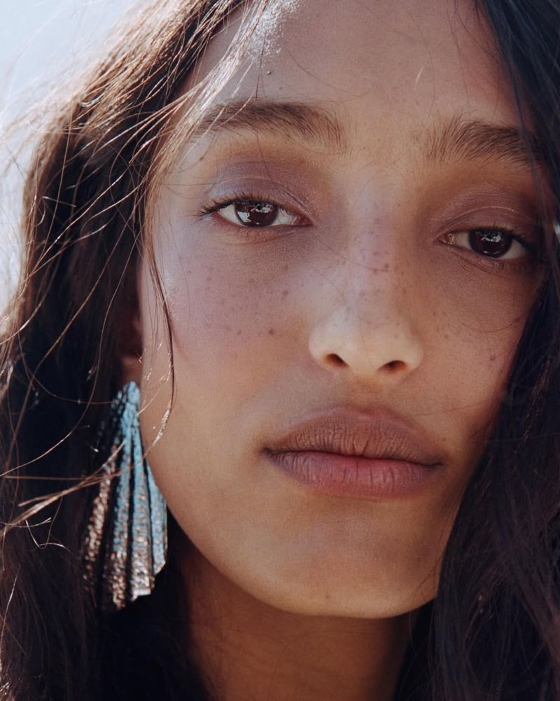 Close-up portrait of a model with dark hair and freckles wearing metallic fan earrings for the H&M Summer 2026 campaign.