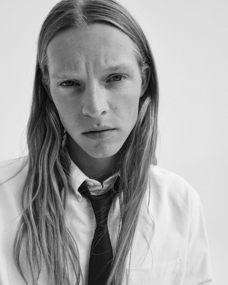 Close-up portrait of a male model with long blonde hair wearing a white shirt and dark tie against a white background
