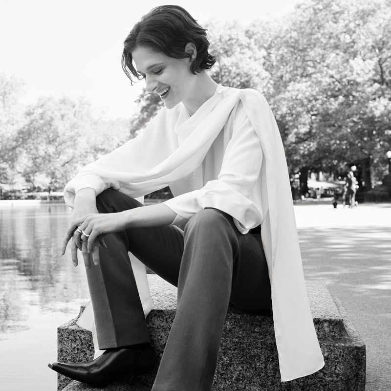 Woman in flowing white top and dark pants sits on a stone ledge near water and trees, smiling in this black-and-white outdoor fashion shot.