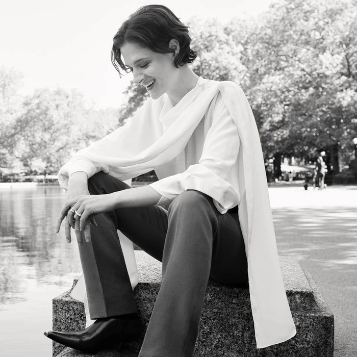 Monochrome portrait of Louisa Jacobson seated outdoors wearing a white draped blouse and dark trousers for American Vogue, photographed by Tess Ayano.