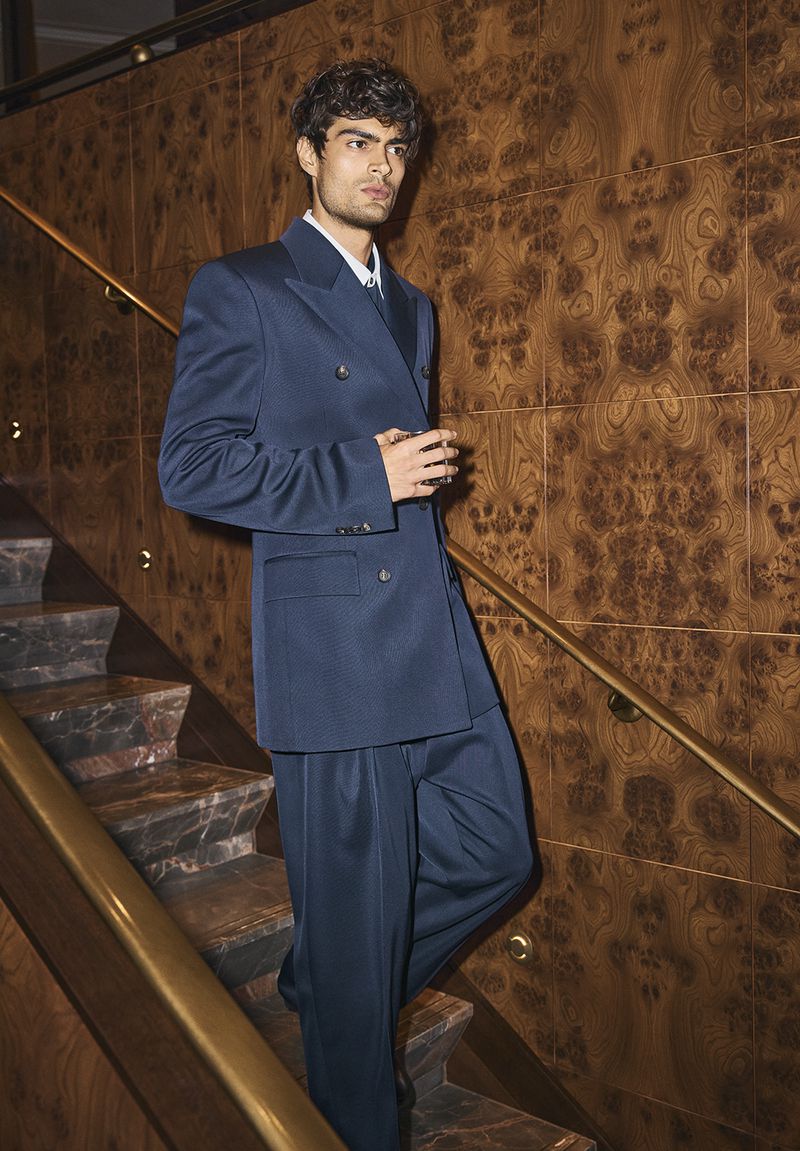 Man wearing a navy blue double-breasted suit descending a staircase with wood-paneled walls.