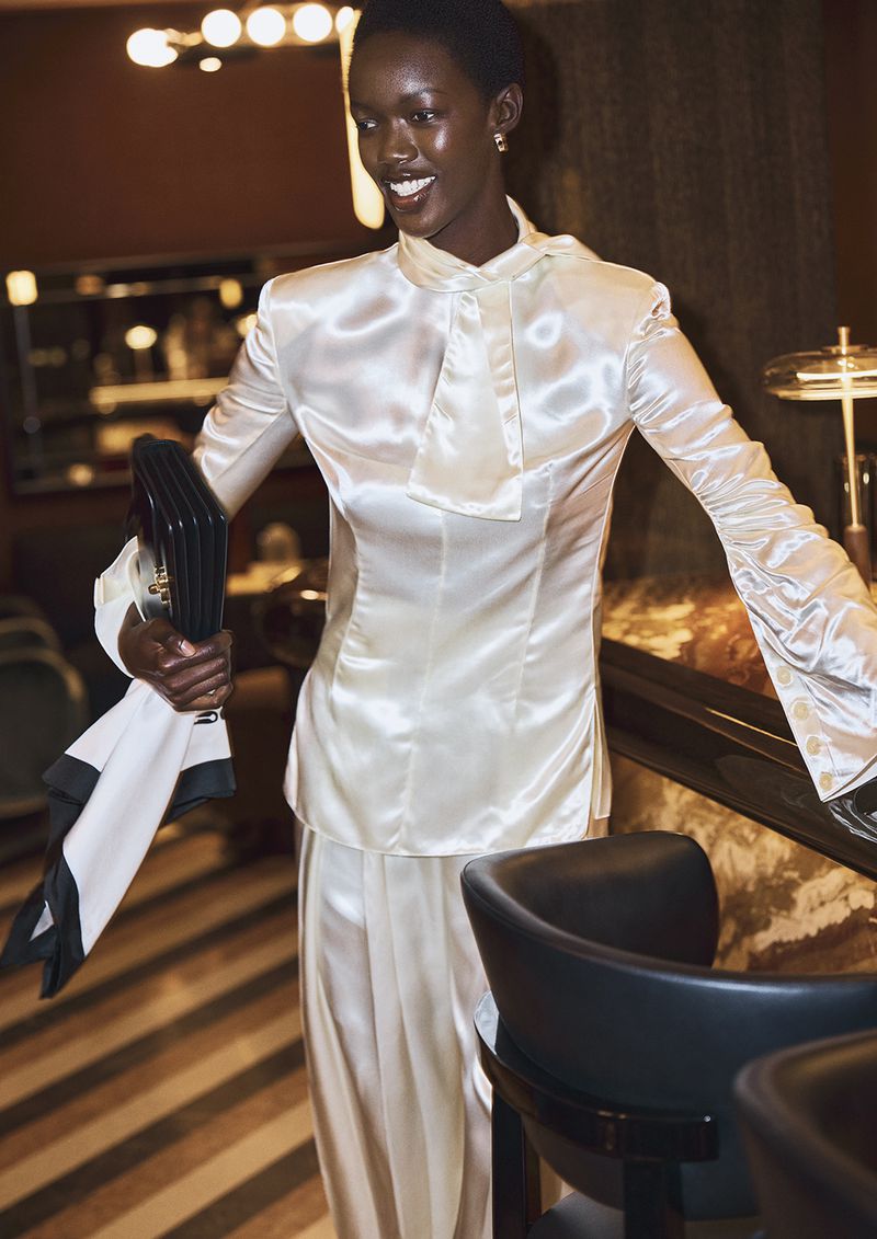 Black woman in a glossy white satin blouse holding a dark handbag in a dimly lit lounge interior.