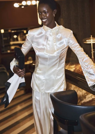 Black woman in a glossy white satin blouse holding a dark handbag in a dimly lit lounge interior.