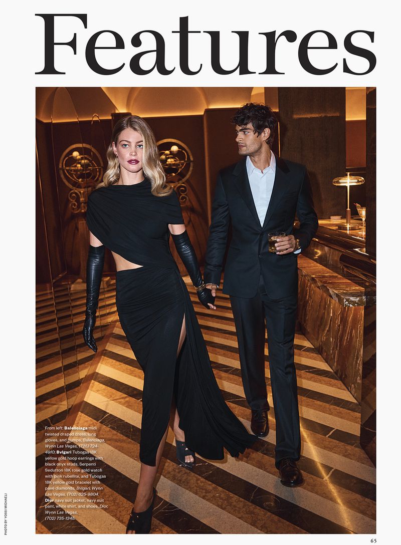 Two models walking through a hotel lobby, wearing black eveningwear and holding hands.