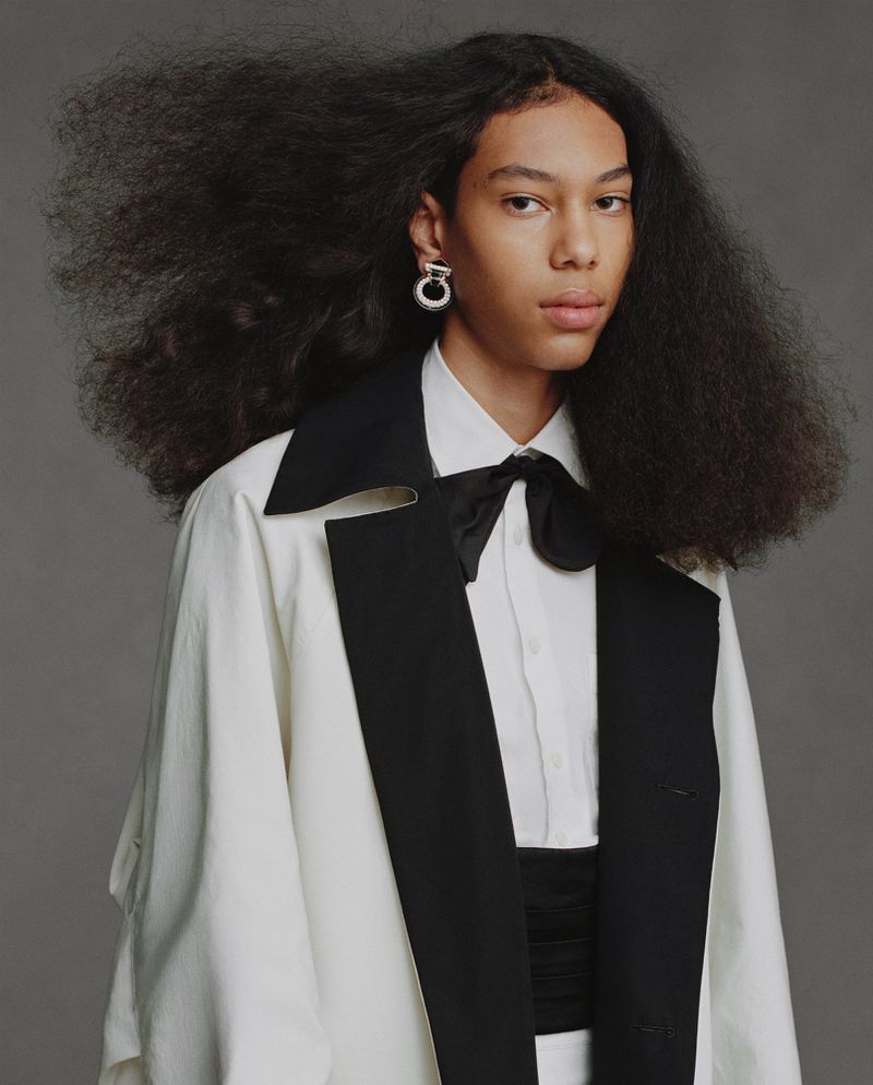 Model with voluminous dark hair wearing a white and black tuxedo jacket, bow tie, and silver earrings against a grey background.