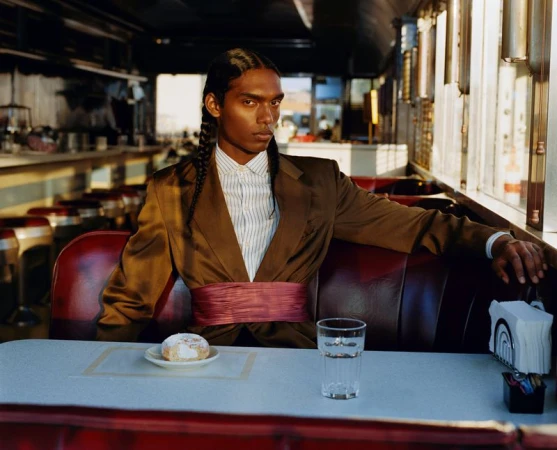 South Asian male model with braided hair seated in a vintage diner booth wearing a brown suit jacket and pink cummerbund.