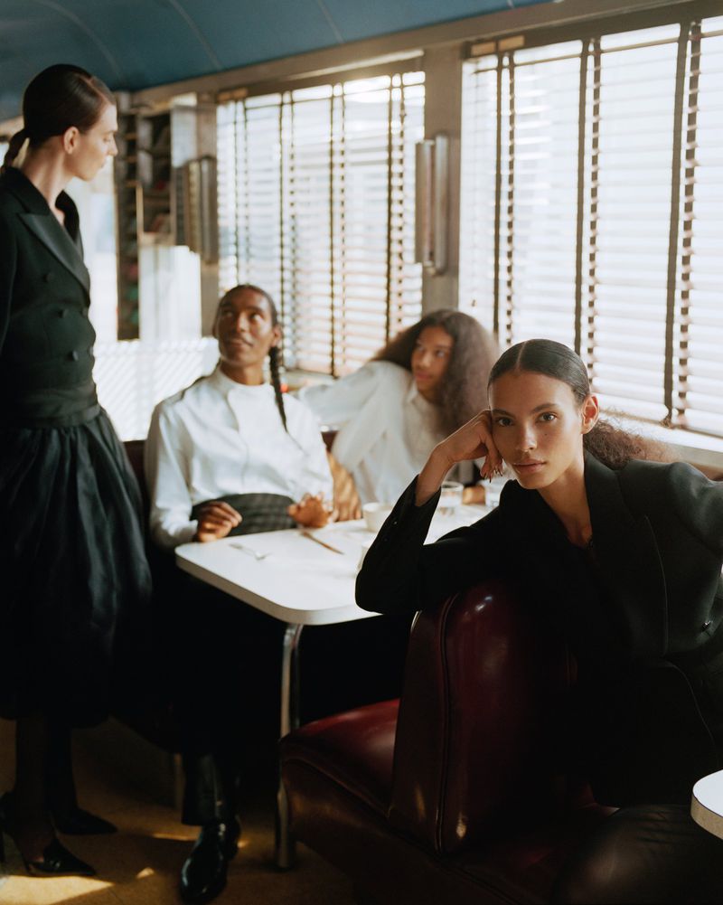 Group of models in formal attire seated and standing inside a vintage diner styled for an editorial shoot by Nadine Ijewere.