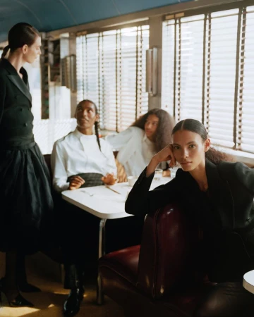 Group of models in formal attire seated and standing inside a vintage diner styled for an editorial shoot by Nadine Ijewere.