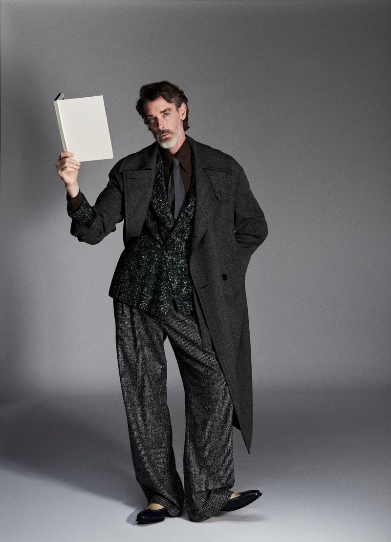Model in charcoal wool coat and patterned suit holding a white book against a grey studio backdrop.