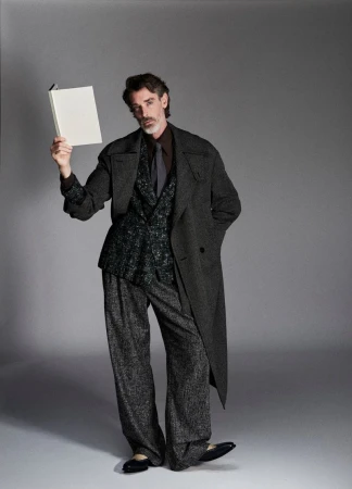 Model in charcoal wool coat and patterned suit holding a white book against a grey studio backdrop.