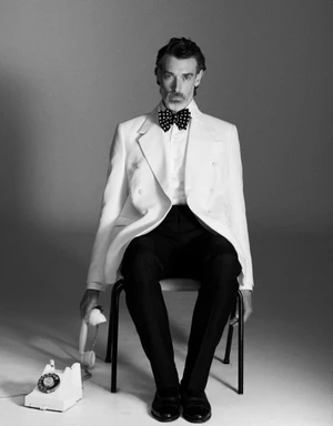 Model Richard Biedul wearing a white double-breasted tuxedo jacket and bow tie, seated on a chair holding a telephone receiver.