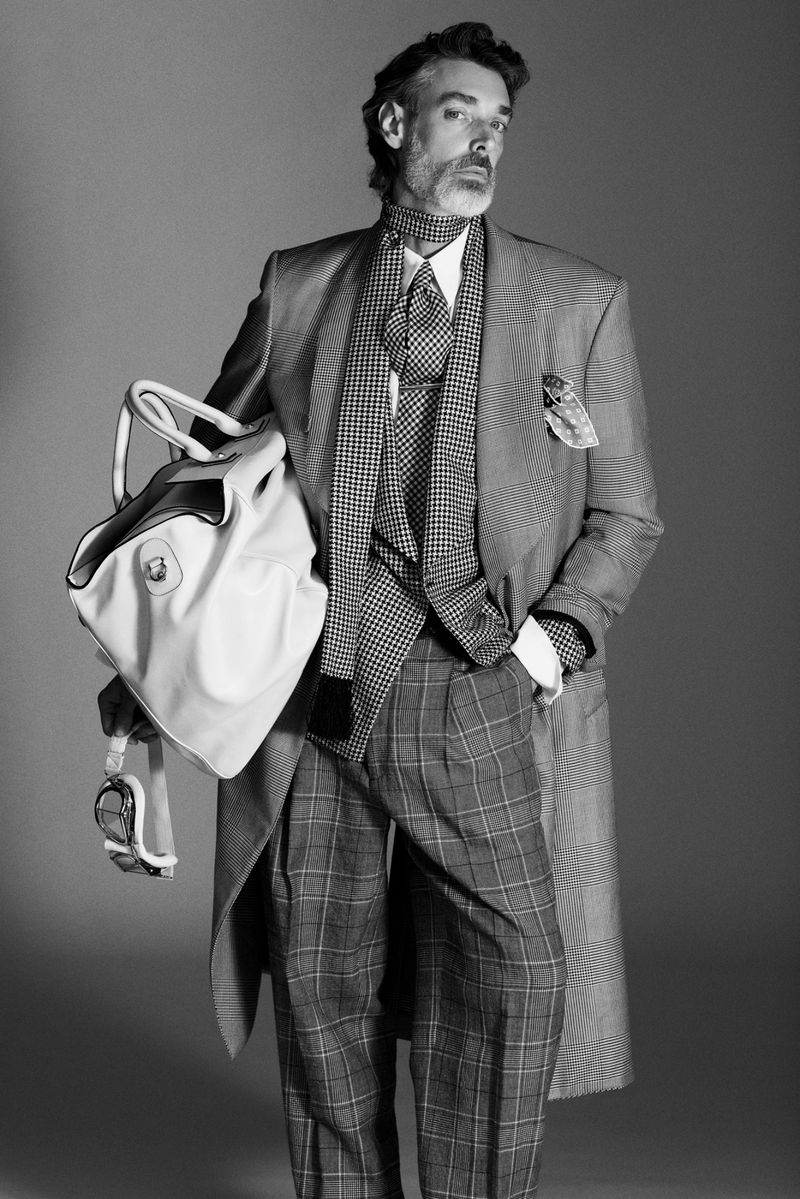 Monochrome portrait of an adult male model wearing a tailored patterned suit holding a large travel bag in a studio.