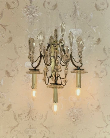An ornate vintage crystal wall sconce with three illuminated candle-style bulbs mounted on patterned wallpaper