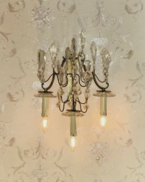 An ornate vintage crystal wall sconce with three illuminated candle-style bulbs mounted on patterned wallpaper